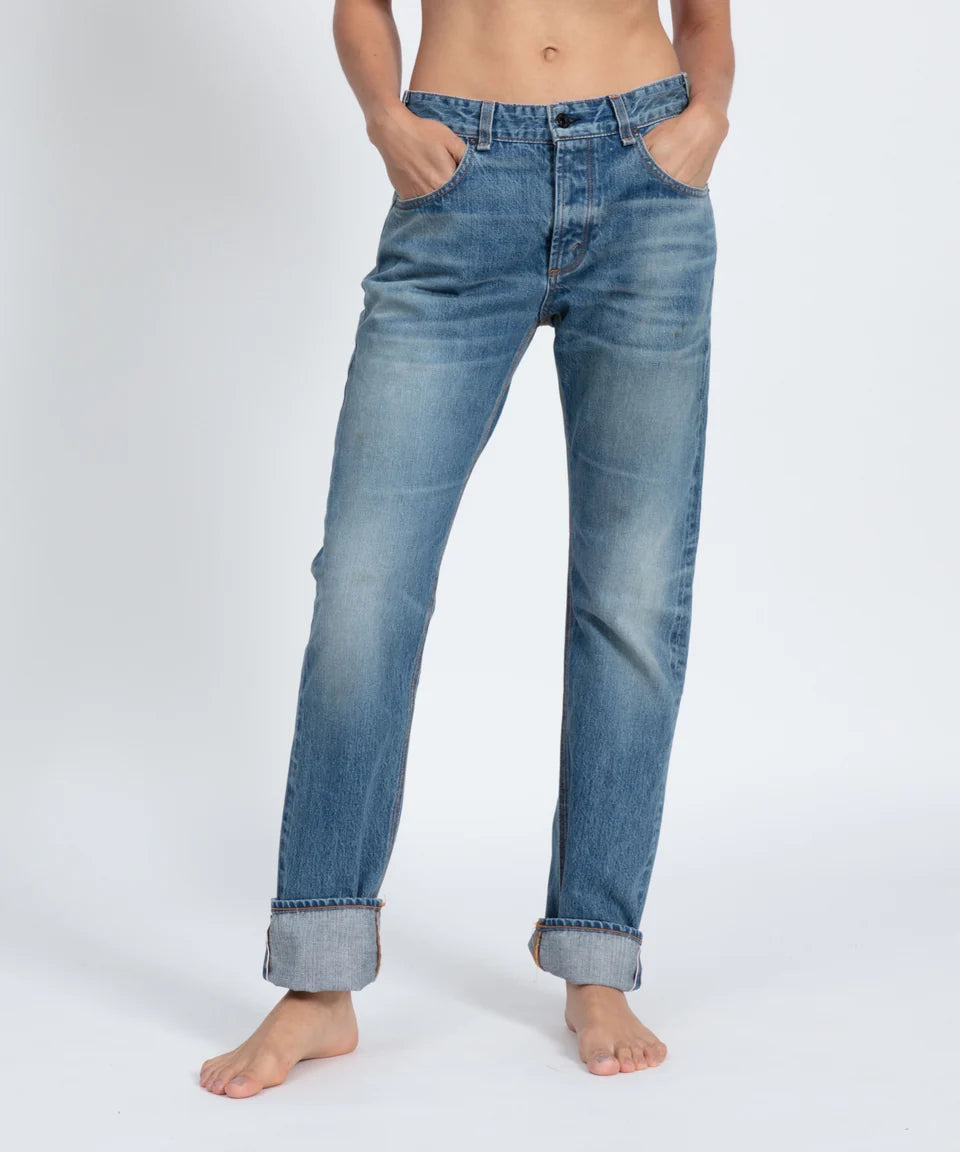 A person stands barefoot against a white background, hands in pockets, wearing ASKK's Selvage Jean. The slightly faded vintage look and relaxed straight leg cut with rolled-up cuffs create a laid-back vibe.