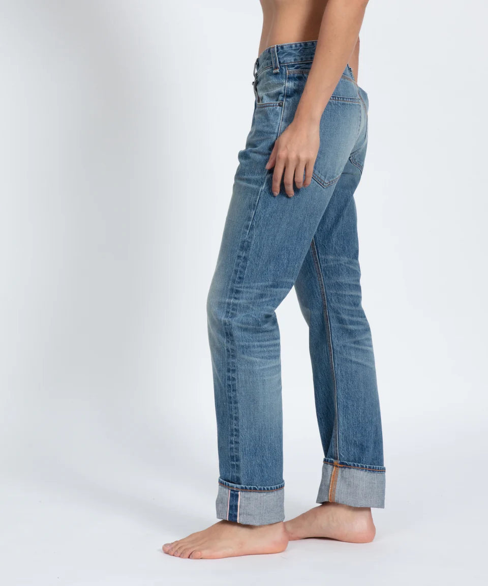 A person stands barefoot on a white surface, wearing ASKK's Selvage Jean in cuffed blue indigo denim with a faded look, visible stitching, and a relaxed fit, shown from waist to ankle in side profile.