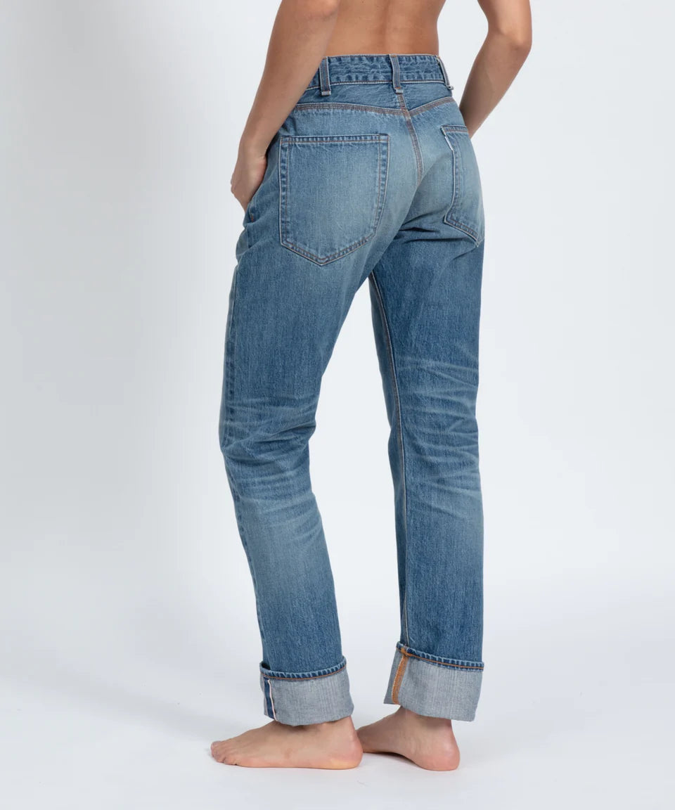 A person stands barefoot from a rear view, wearing ASKK's Selvage Jean—a pair of blue, cuffed straight-leg jeans made of indigo selvage denim. The vintage men's jeans feature visible stitching and back pockets against a light background, with the upper body not fully visible.