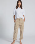 A woman stands barefoot against a plain backdrop, wearing a loose-fitting white T-shirt and ASKK's beige rolled-up chino twill pants. Her hands are in her pockets, and she is looking directly at the camera with a neutral expression.