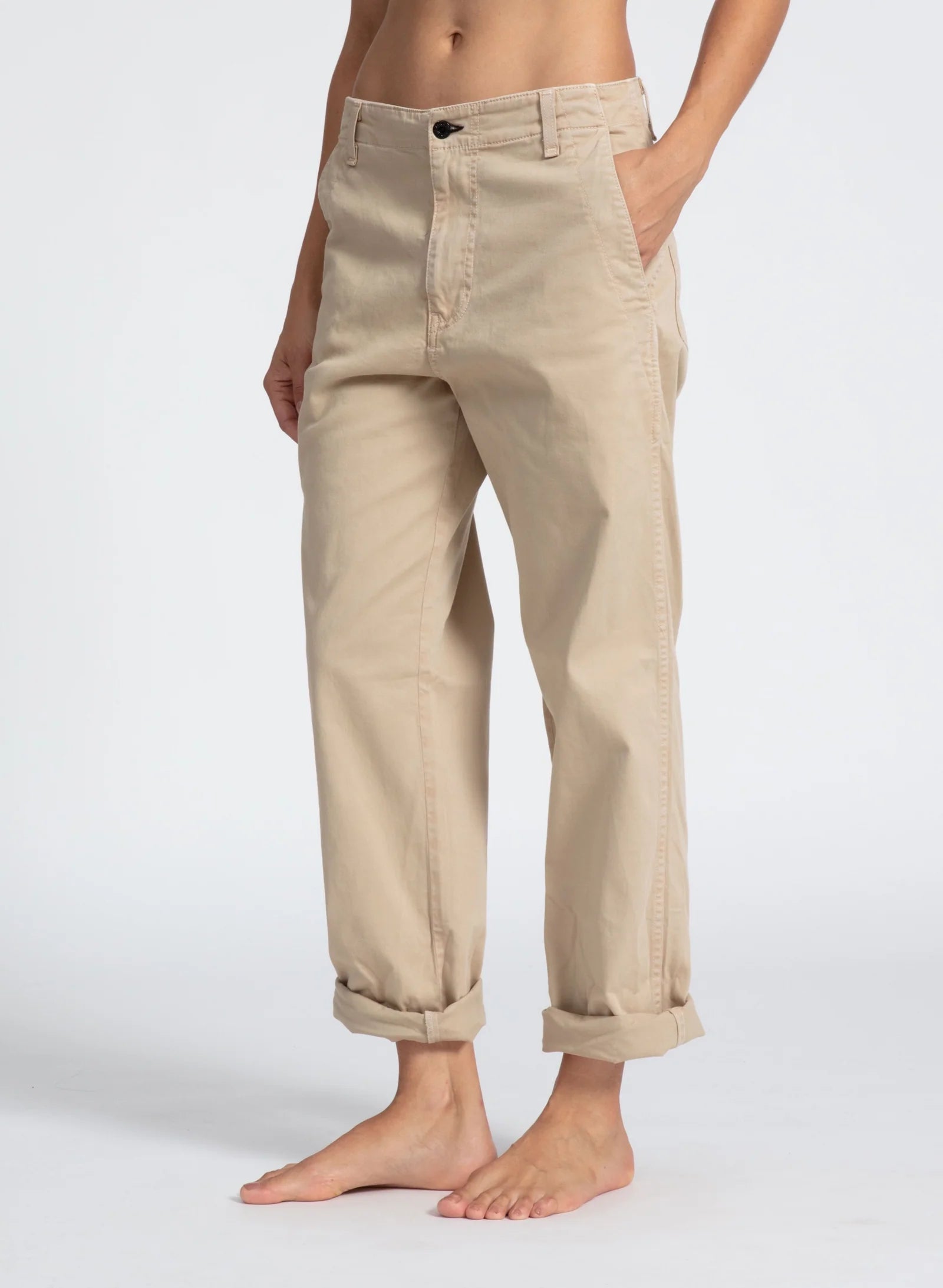 A person is standing with their hands in the pockets of ASKK chino twill pants in beige. The relaxed chinos are rolled up at the cuffs, and they are barefoot. The background is plain and neutral, highlighting the pants. The person's head and upper torso are not visible.