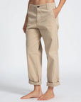 A person is standing with their hands in the pockets of ASKK chino twill pants in beige. The relaxed chinos are rolled up at the cuffs, and they are barefoot. The background is plain and neutral, highlighting the pants. The person's head and upper torso are not visible.