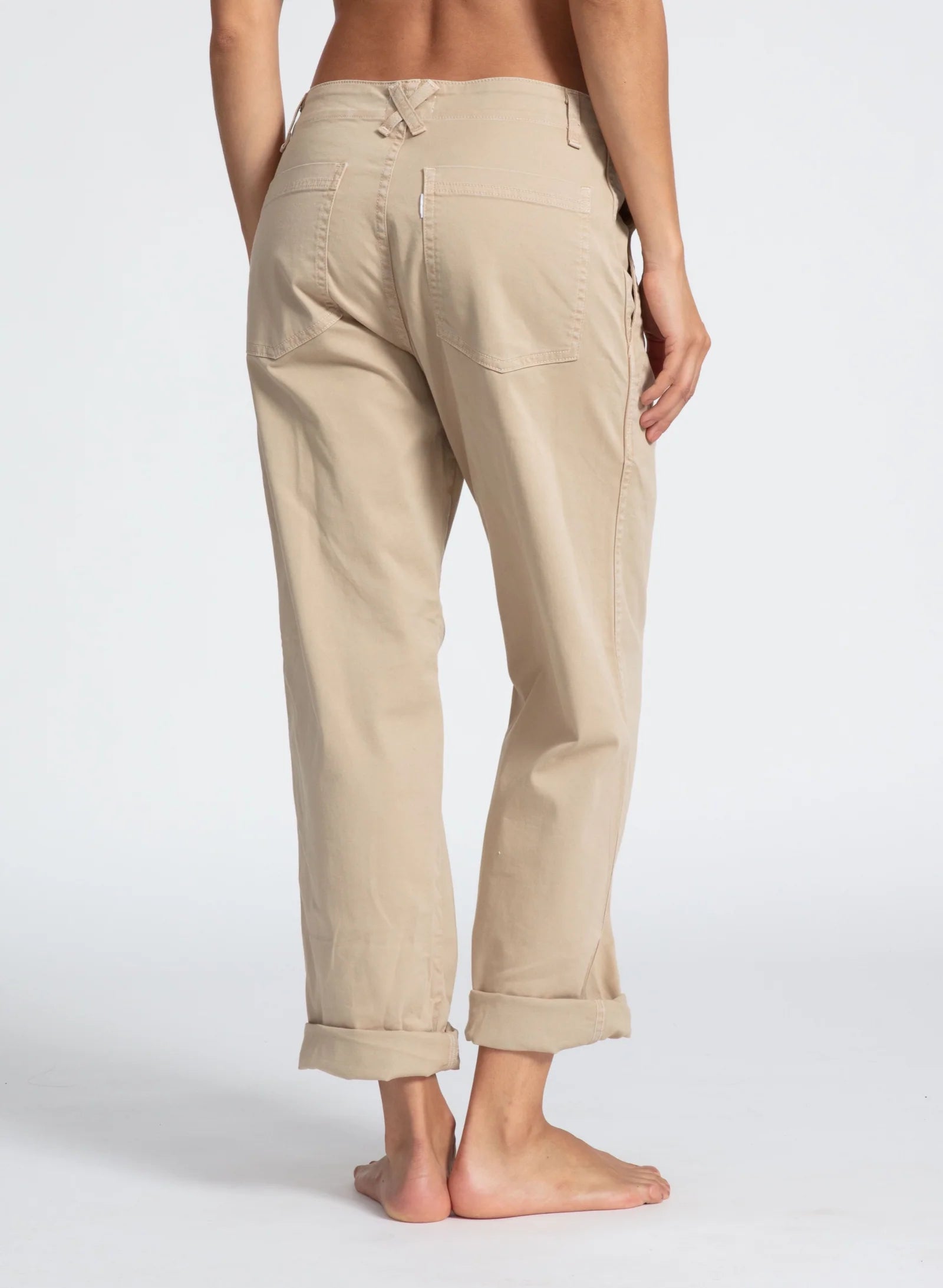 A person stands barefoot with their back to the camera, showcasing ASKK's light beige, rolled-up chino twill pants. These relaxed-fit pants feature visible back pockets and taper slightly at the ankles against a plain white background.