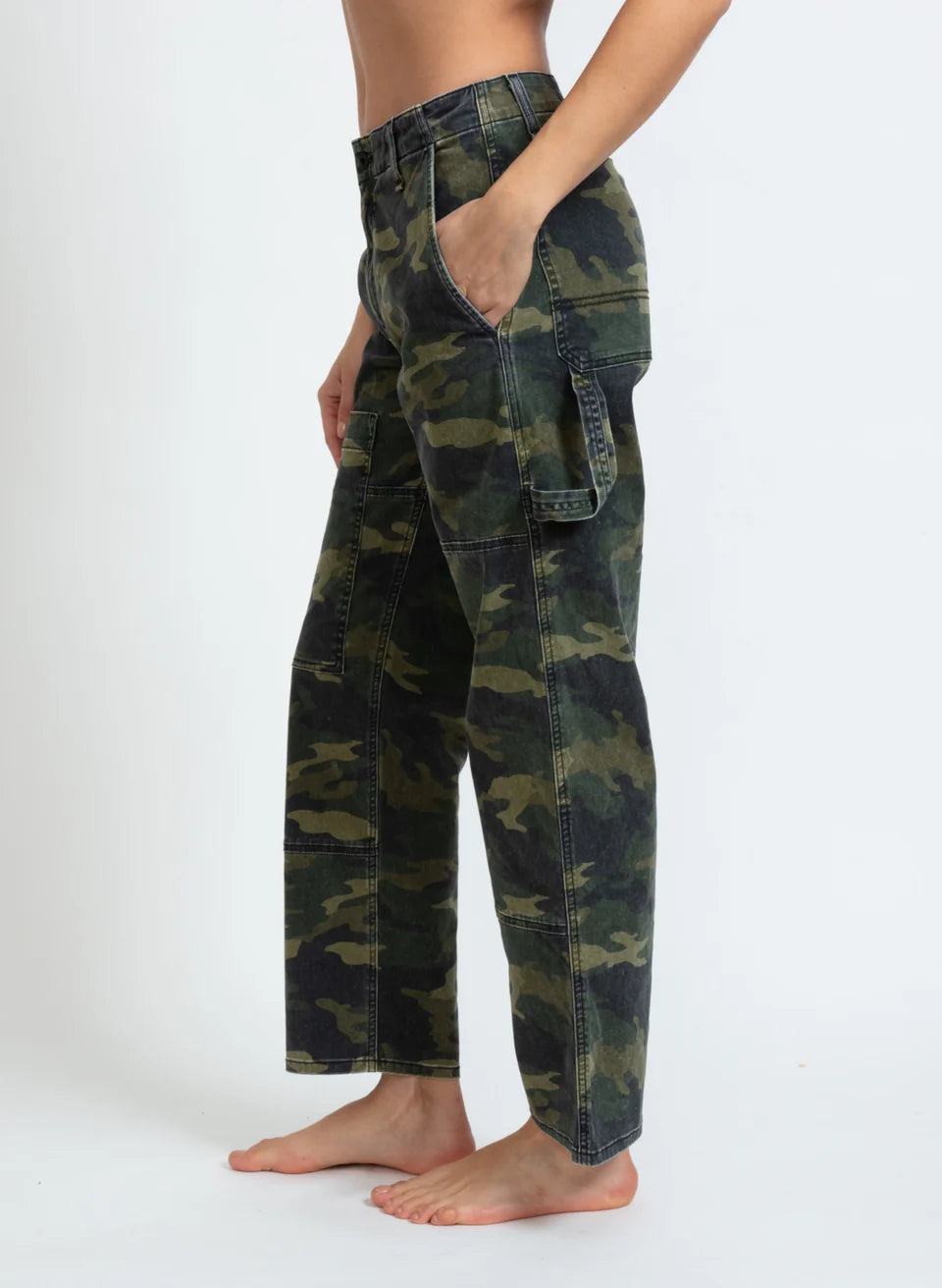 A person stands barefoot against a plain background, wearing ASKK's Carpenter pants in green camouflage. These stretch twill cargo pants, crafted with large side pockets and a relaxed fit, are made in Los Angeles. Their hands rest comfortably in the pockets.