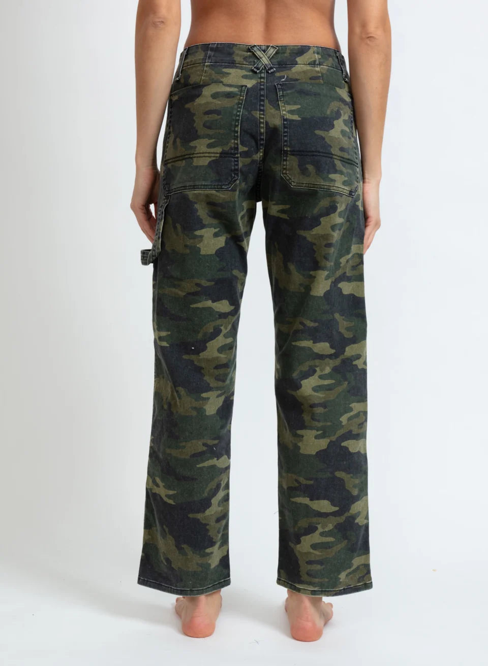 A person is pictured standing barefoot, highlighting the rear view of ASKK's Carpenter pants in green camouflage. These relaxed-fit cargo pants are constructed from stretch twill and boast large pockets along with the rugged appeal typical of carpenter-style attire. Made in Los Angeles, they make a striking contrast against a simple white backdrop.