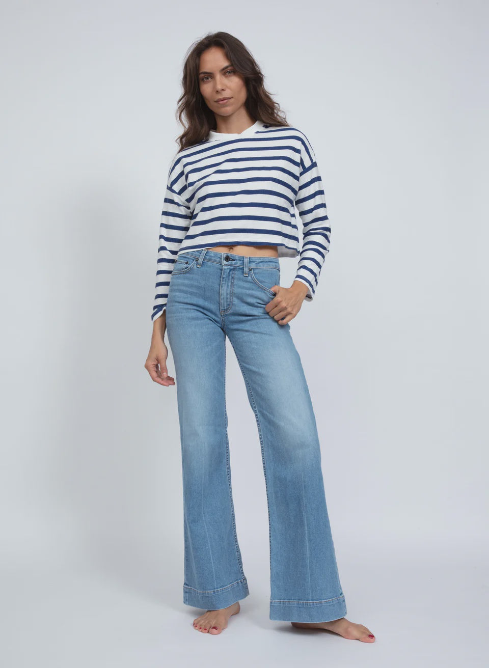 A person with long hair poses confidently in an ASKK Cropped LS Tee, a white and navy striped shirt made from American cotton, paired with blue bell-bottom jeans. They stand barefoot against a plain white background, exuding effortless style.