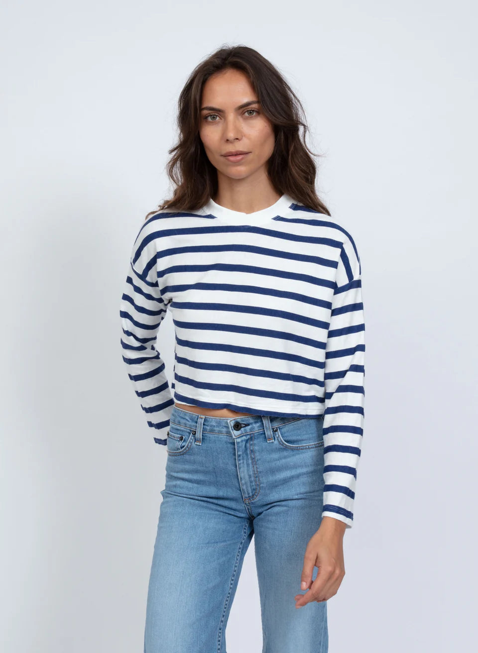 A person with long brown hair wears the ASKK Cropped LS Tee, featuring a white and blue stripe design. Paired with blue jeans, they stand against a plain white backdrop with a neutral expression, capturing the casual elegance of Los Angeles fashion.