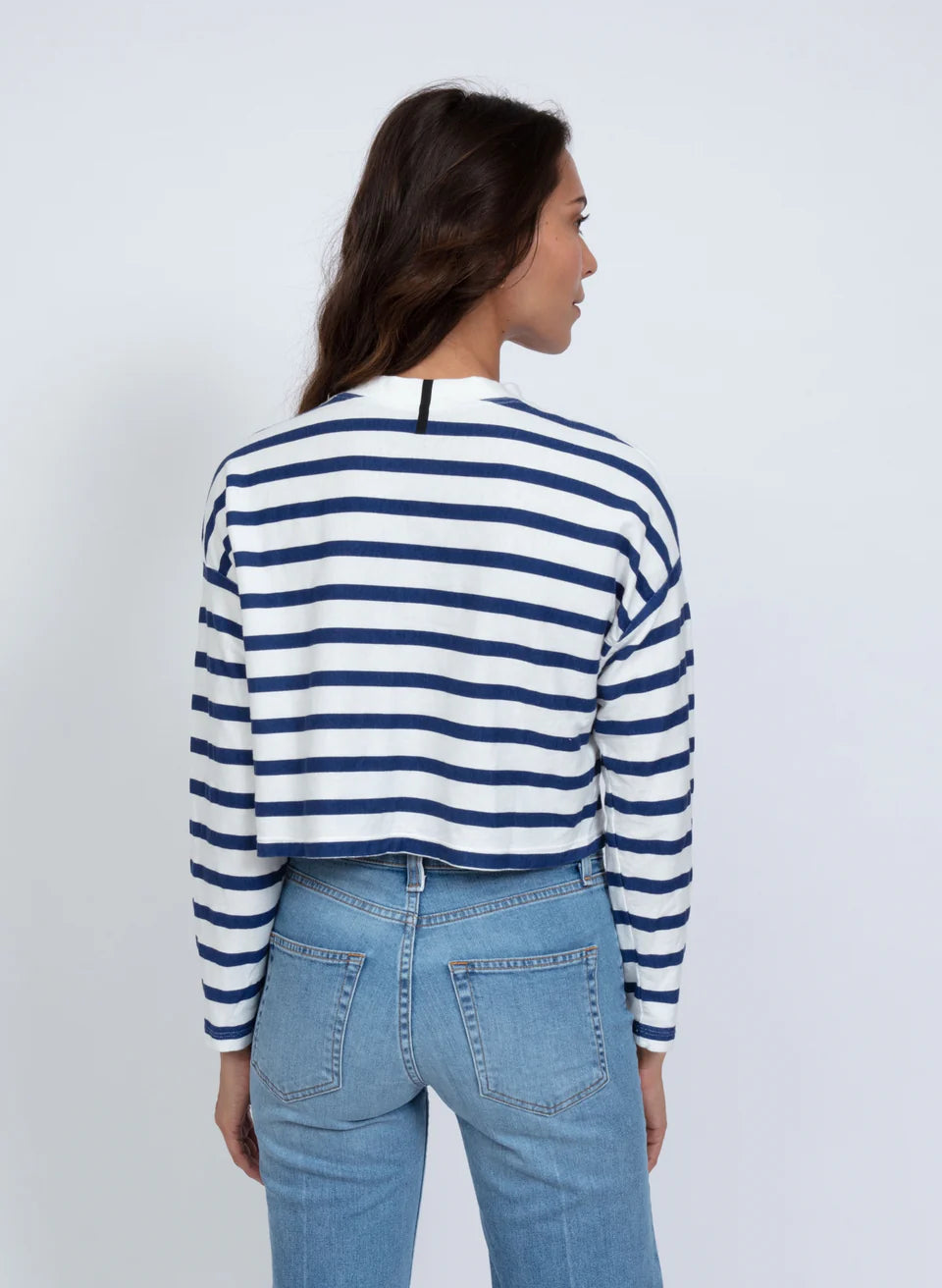 A person with long hair wears ASKK’s Cropped LS Tee, a blue and white striped long-sleeve top, paired with light blue jeans. Made in Los Angeles from American fabric, they stand away from the camera against a plain white background.