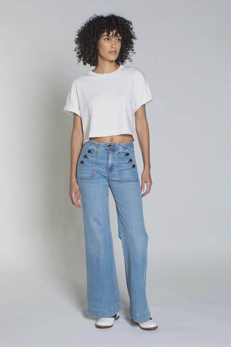 A person with curly hair stands against a plain background, dressed in an ASKK white cotton Cuff Tee and light blue wide-legged jeans featuring decorative buttons—a nod to classic American fabric. They complete the Los Angeles-inspired look with stylish white shoes.