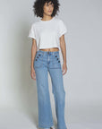 A person with curly hair stands against a plain background, dressed in an ASKK white cotton Cuff Tee and light blue wide-legged jeans featuring decorative buttons—a nod to classic American fabric. They complete the Los Angeles-inspired look with stylish white shoes.