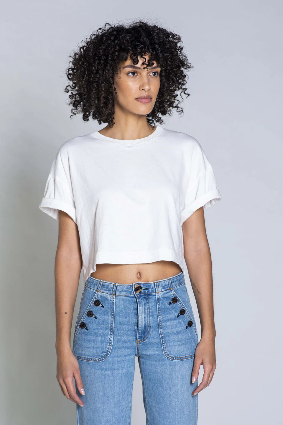 With her curly hair, she wears ASKK's Cuff Tee, a white cropped cotton t-shirt, paired with high-waisted blue jeans detailed with small black buttons from American fabric. Posed against a plain gray background, this Los Angeles-crafted ensemble effortlessly merges style and comfort.