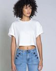 With her curly hair, she wears ASKK's Cuff Tee, a white cropped cotton t-shirt, paired with high-waisted blue jeans detailed with small black buttons from American fabric. Posed against a plain gray background, this Los Angeles-crafted ensemble effortlessly merges style and comfort.