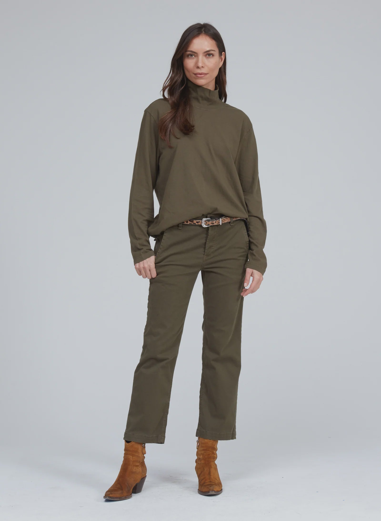 A woman stands confidently wearing an olive-green long-sleeve turtleneck and a matching pair of ASKK&#39;s Slim Chino. She accessorizes with a belt and brown ankle boots. Her long dark hair is styled loosely, and she poses in front of a plain, light gray background.