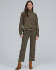 A woman stands confidently wearing an olive-green long-sleeve turtleneck and a matching pair of ASKK's Slim Chino. She accessorizes with a belt and brown ankle boots. Her long dark hair is styled loosely, and she poses in front of a plain, light gray background.