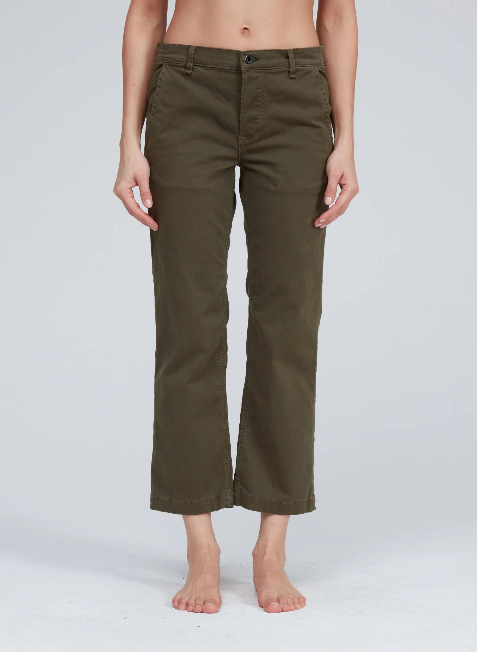 A person with bare feet is wearing olive green Slim Chino pants from ASKK. The pants feature a button and zipper closure, front pockets, and offer a tailored fit. Made in Los Angeles, the background is plain and light-colored.