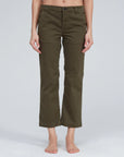 A person with bare feet is wearing olive green Slim Chino pants from ASKK. The pants feature a button and zipper closure, front pockets, and offer a tailored fit. Made in Los Angeles, the background is plain and light-colored.