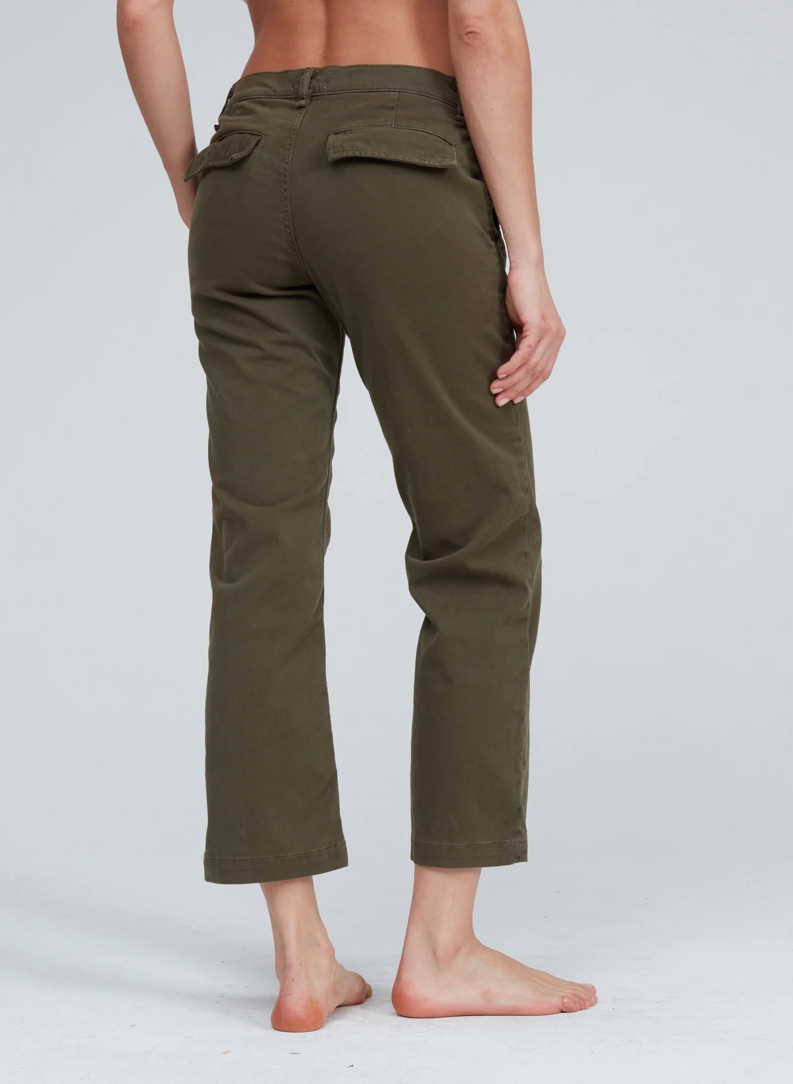 A person stands barefoot with their back to the camera, wearing ASKK&#39;s Slim Chino pants in olive green with pockets. The background is plain and light gray.