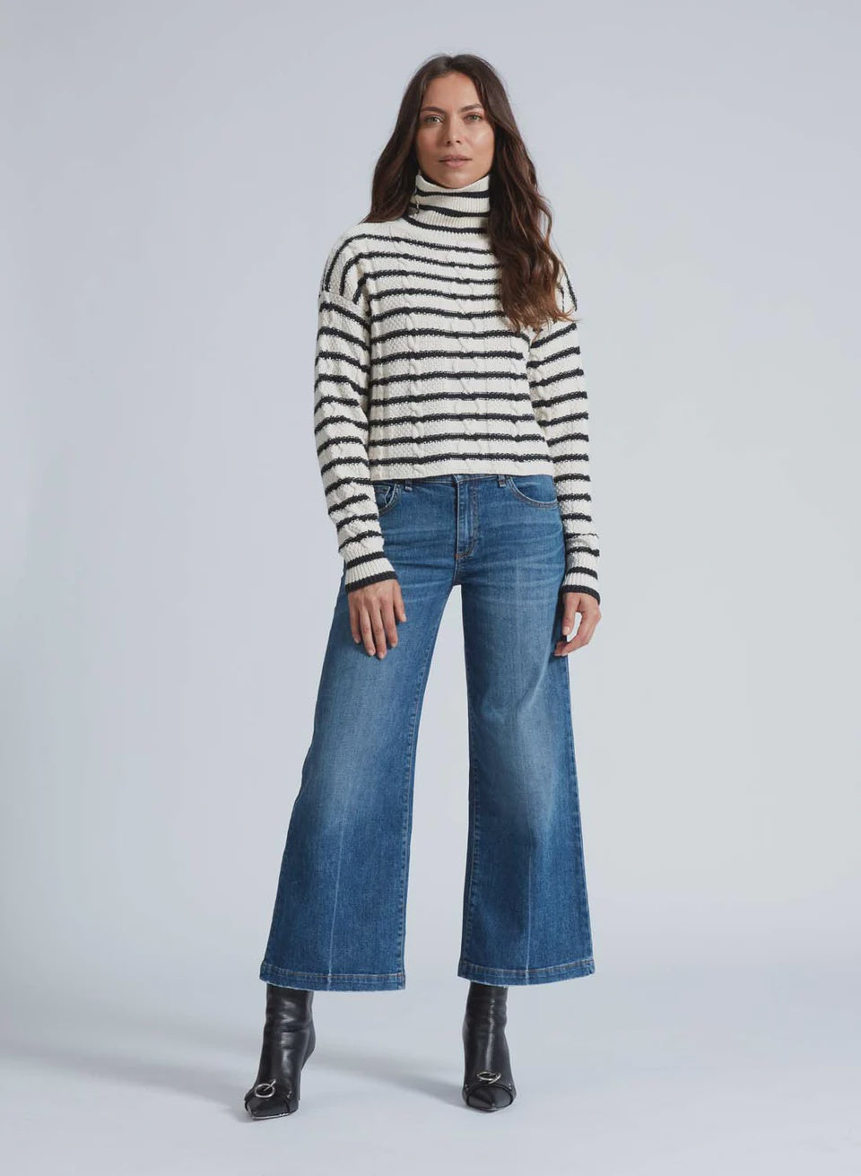 A person with long hair is wearing the black and white striped Cable Crop Stripe sweater by ASKK, paired with wide-leg blue jeans and black ankle boots. They stand against a plain background.