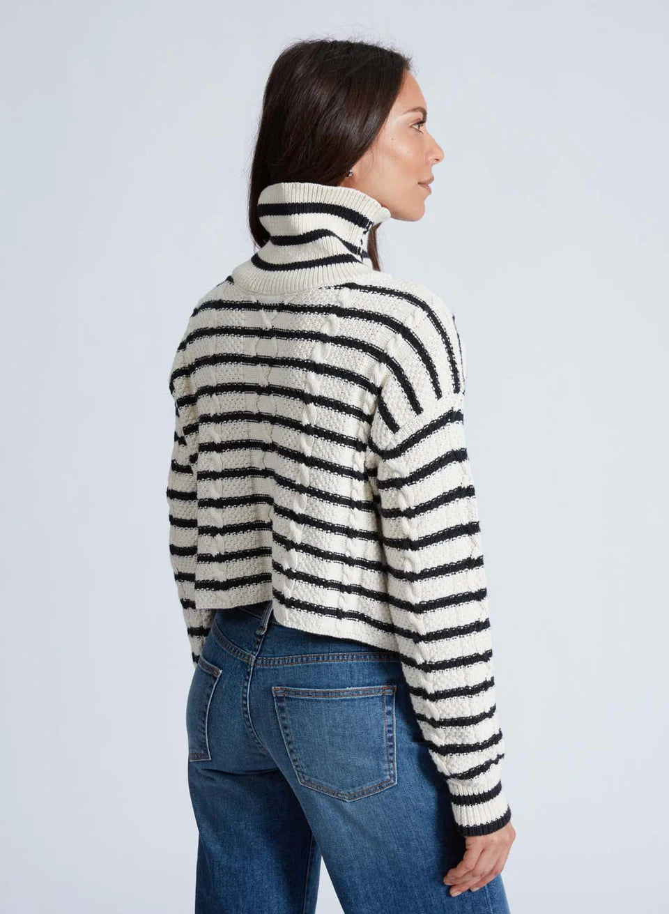 A person with long hair is elegantly dressed in ASKK's Cable Crop Stripe, a chic black and white striped cropped turtleneck sweater crafted from organic cotton. They pair it effortlessly with blue jeans and stand gracefully against a plain background, gazing thoughtfully to the side.