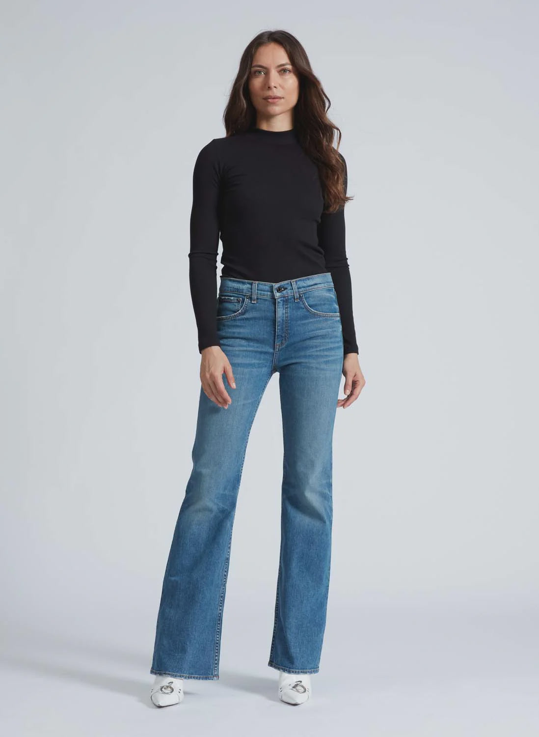 A woman with long brown hair wears the ASKK Mock Neck Long Sleeve, a fitted black ribbed top, paired with high-waisted blue jeans that flare at the bottom. She stands against a plain light grey background, looking at the camera with a neutral expression, hands relaxed at her sides.