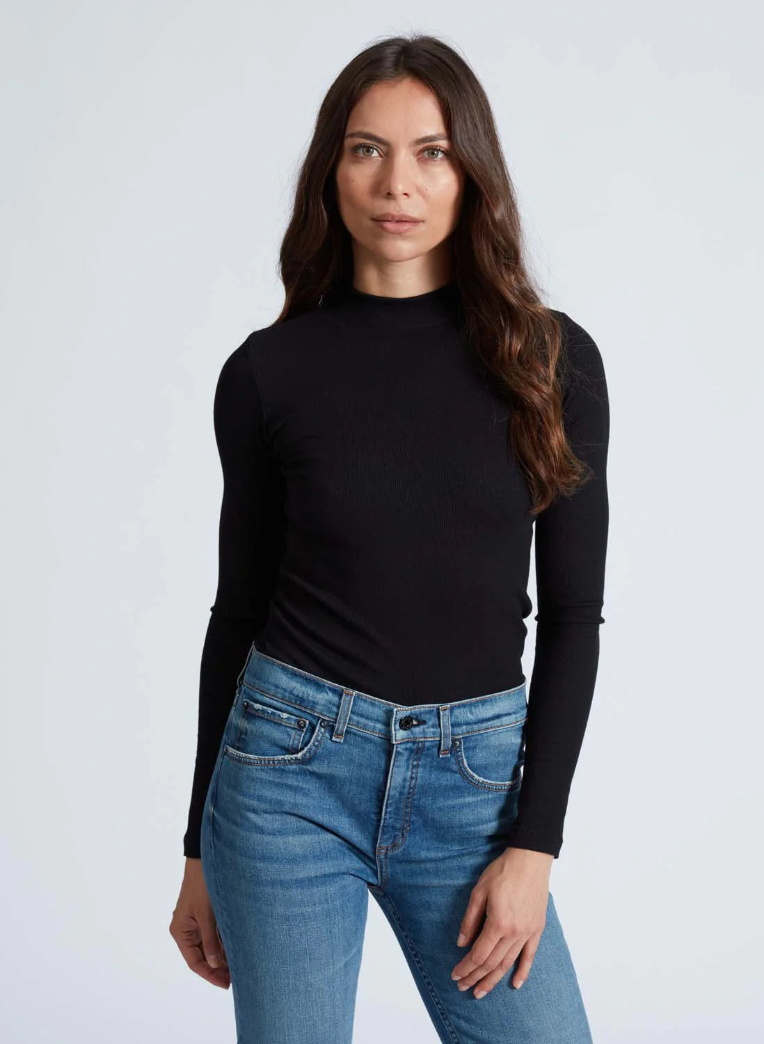 A woman with long, wavy brown hair is wearing an ASKK Mock Neck Long Sleeve top paired with blue jeans. She is standing against a plain white background, looking directly at the camera with a neutral expression.