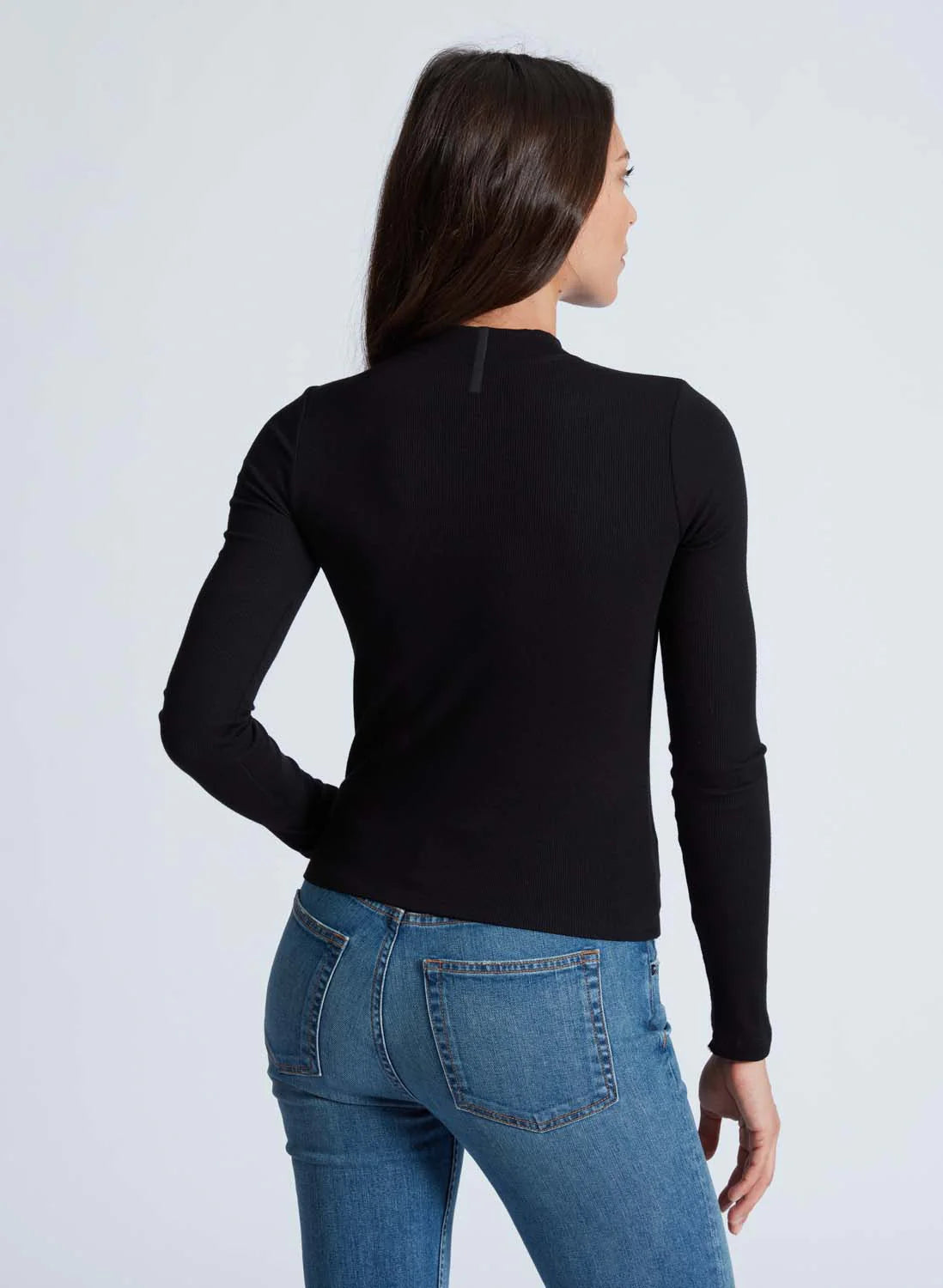 A woman with long, dark hair is seen from behind, wearing an ASKK Mock Neck Long Sleeve top and blue jeans. She stands against a plain, light background, her right hand resting on her hip and her left arm hanging down by her side.