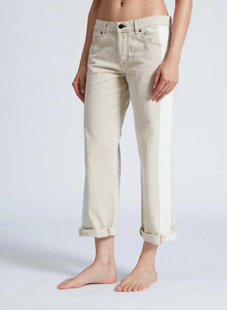 A person stands barefoot in ASKK's Factory Girl jeans, featuring low-rise, light beige Japanese denim with rolled cuffs and a white side stripe. Crafted in Los Angeles, they're set against a plain, light-colored background.