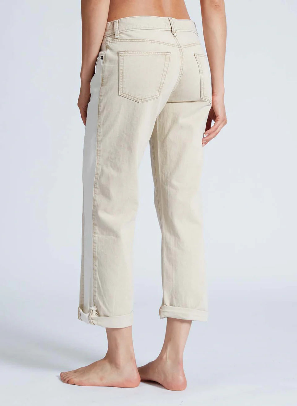 A person stands barefoot in low-rise, light beige rolled-up jeans named Factory Girl from ASKK, made of premium Japanese denim. The visible back pockets enhance the casual and relaxed vibe against a plain background.