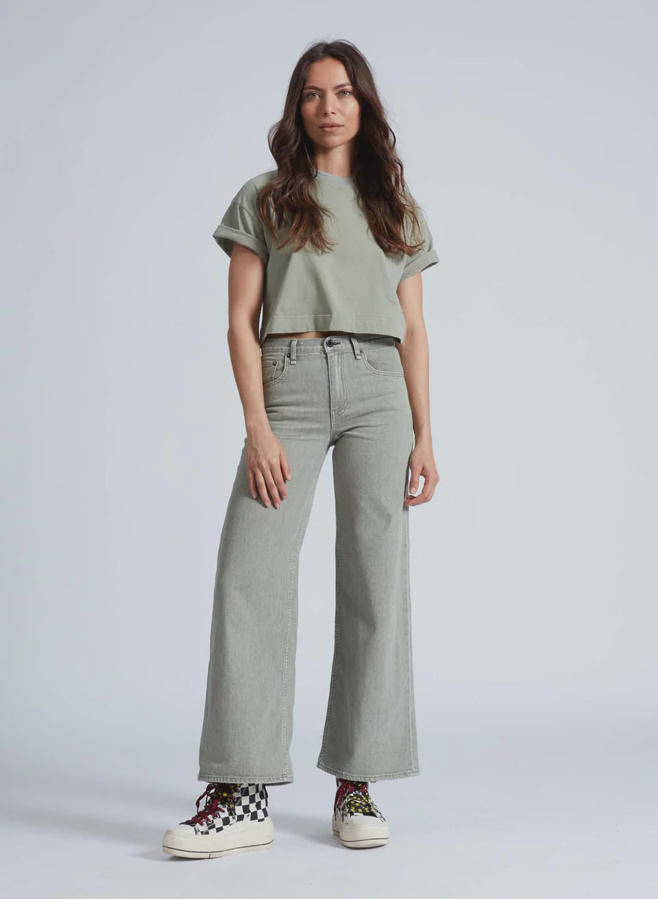 Standing against a gray background, a person with long hair wears an ASKK light green T-shirt and stylish Japanese denim Slim Wide Leg jeans. Colorful high-tops with black and white checkered patterns complete the look, blending urban flair from Los Angeles.