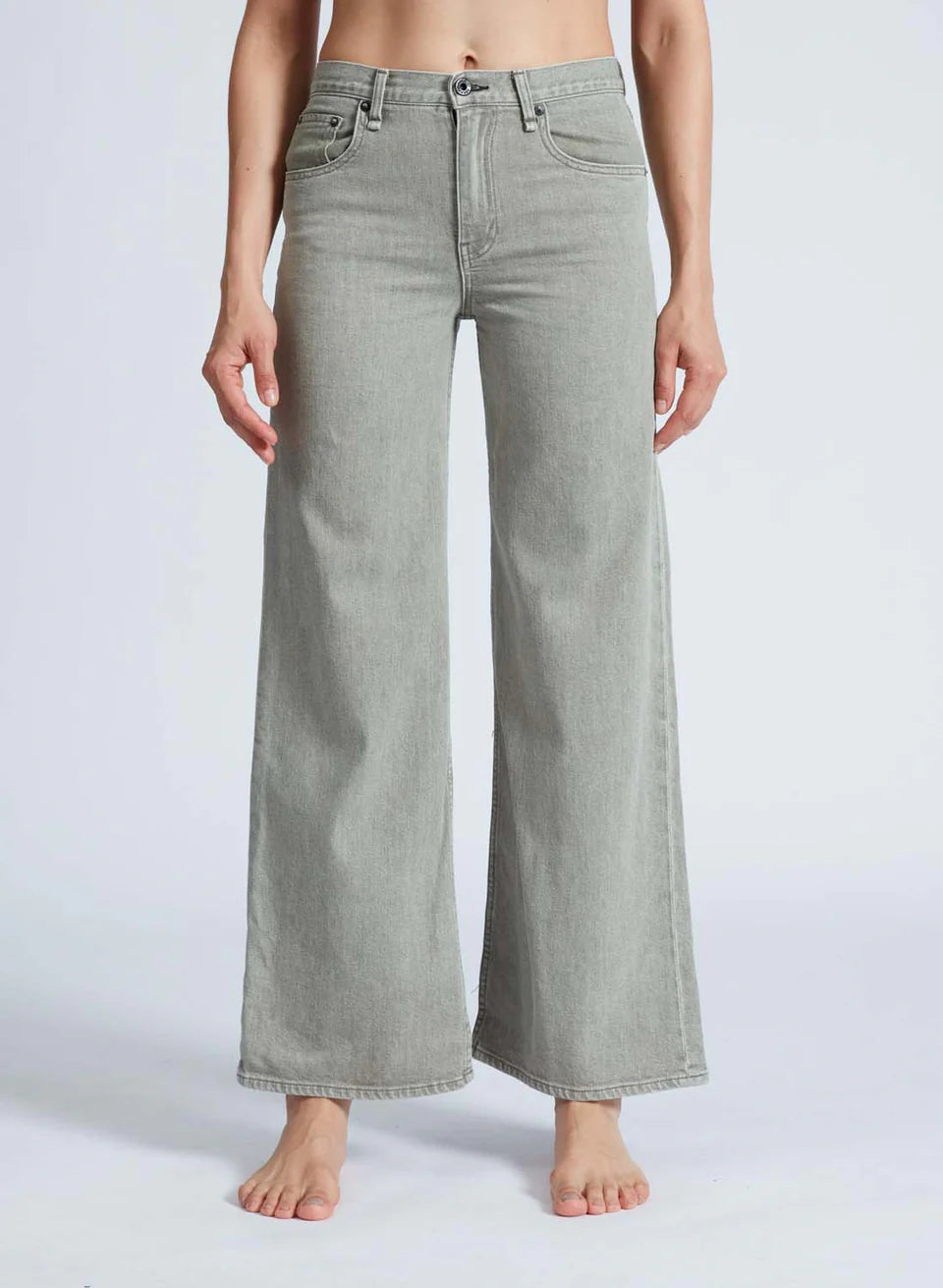 Barefoot against a plain background, a person dons ASKK's Slim Wide Leg jeans. Tailored in Los Angeles with Japanese denim, these light gray, high-waisted jeans feature front pockets and a button closure.