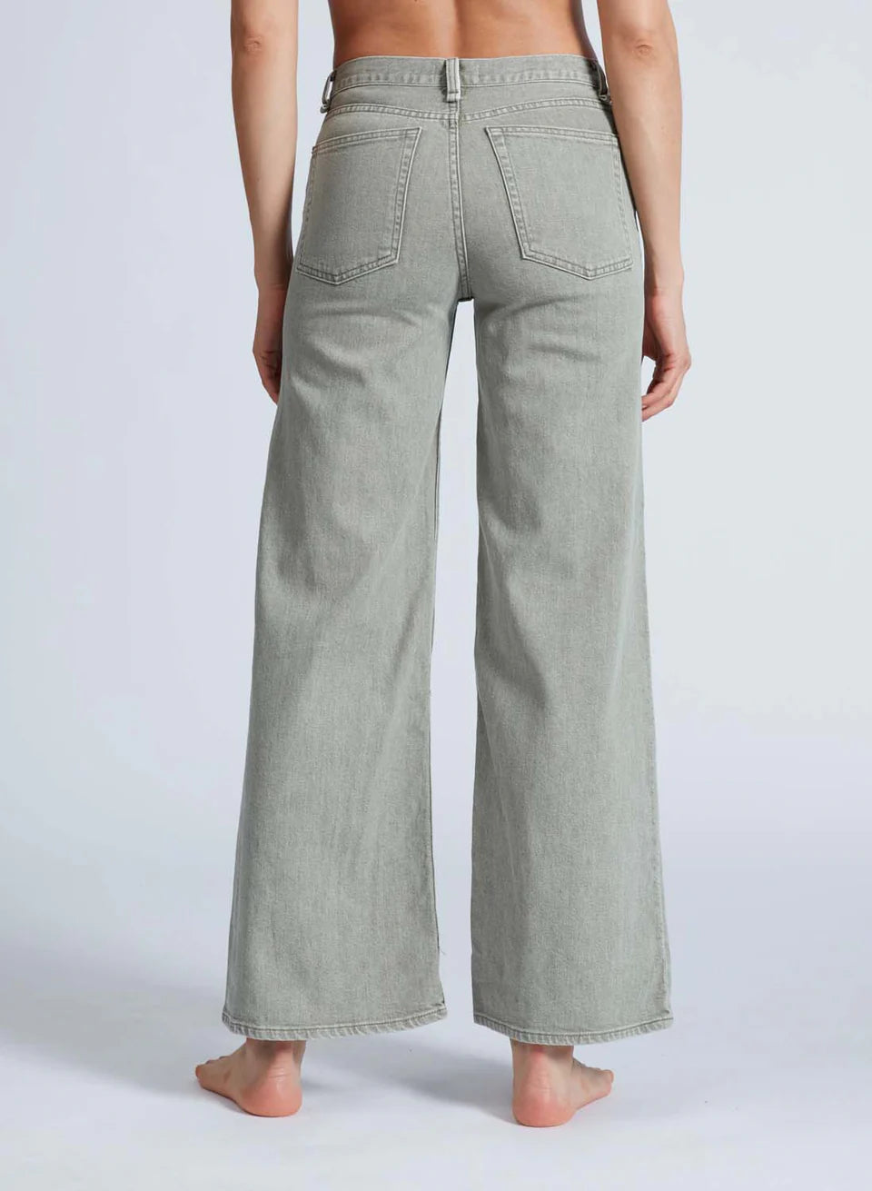 A barefoot person stands on a light background, back to the camera, wearing ASKK's high-waisted Slim Wide Leg gray jeans. Made from Japanese denim, these jeans feature two back pockets and a loose fit.