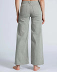 A barefoot person stands on a light background, back to the camera, wearing ASKK's high-waisted Slim Wide Leg gray jeans. Made from Japanese denim, these jeans feature two back pockets and a loose fit.