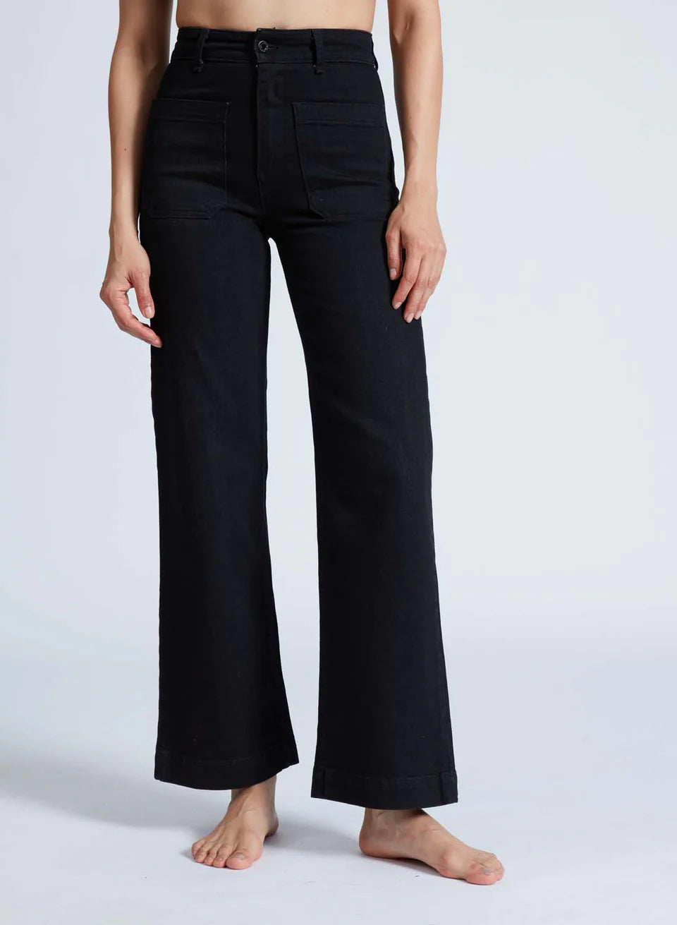 A person is modeling the ASKK Sailor Pant, which are high-rise, black wide-leg pants crafted from Japanese denim. They stand barefoot against a plain white background. The pants feature a tailored fit around the hips and a loose fit from the thighs downward.