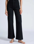 A person is modeling the ASKK Sailor Pant, which are high-rise, black wide-leg pants crafted from Japanese denim. They stand barefoot against a plain white background. The pants feature a tailored fit around the hips and a loose fit from the thighs downward.