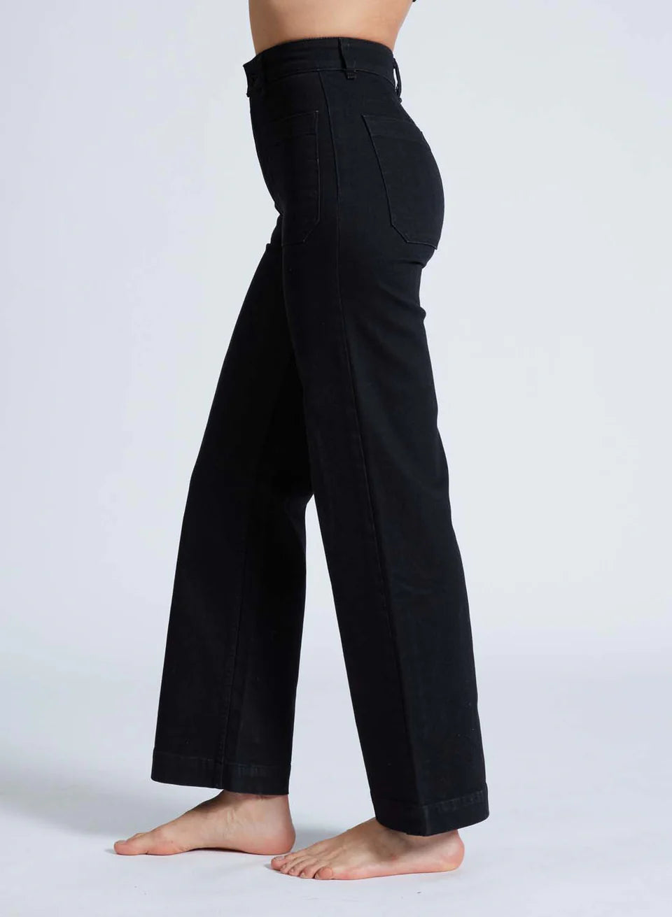 A person is standing barefoot on a light gray surface, wearing ASKK's Sailor Pant. These high-waisted, wide-leg pants are black with visible seams and crafted from Japanese denim, leaving their upper body out of the frame.