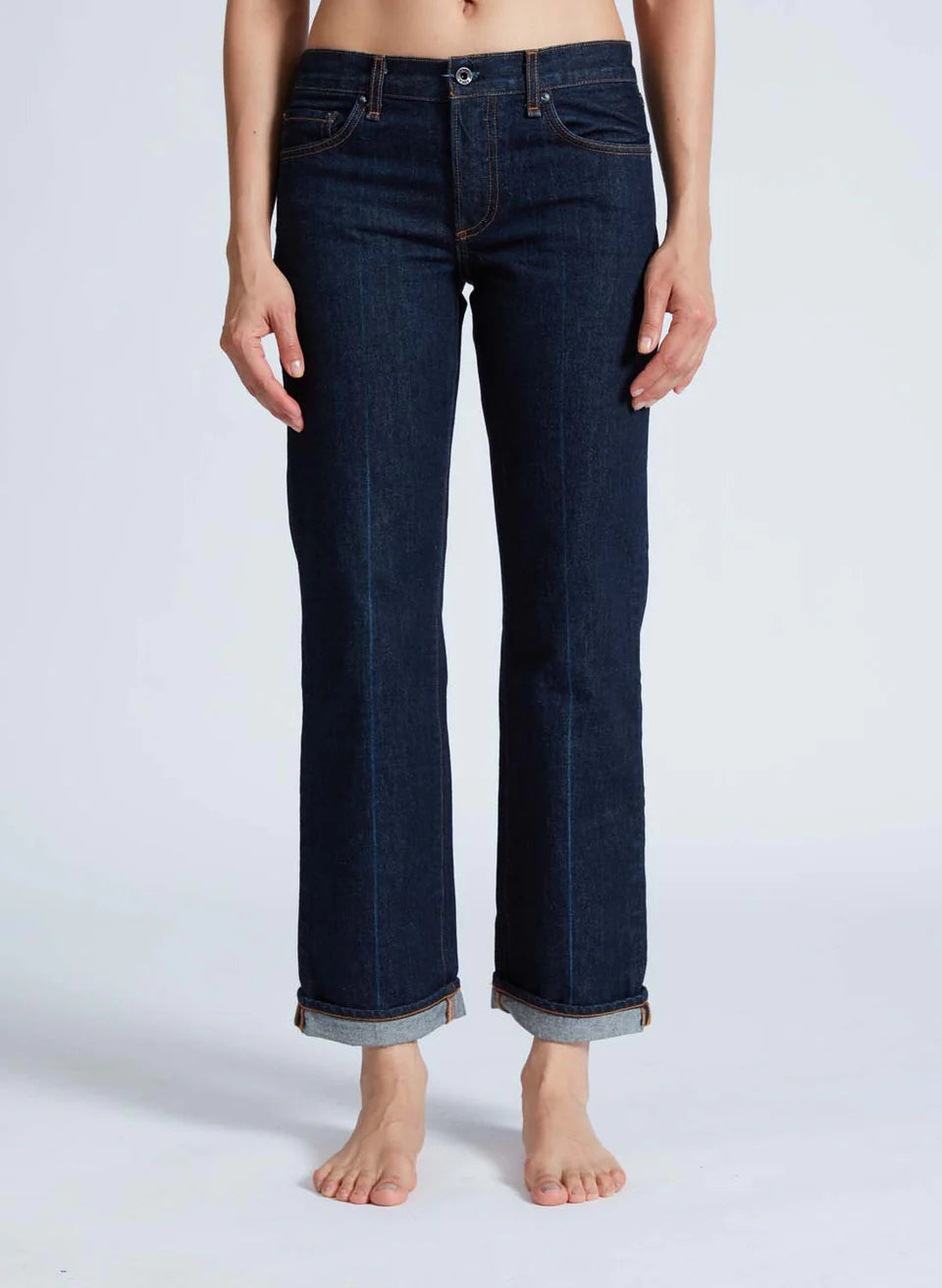 An individual is barefoot against a plain white background, wearing the ASKK Low Rise Straight jeans in dark blue, which have a rolled cuff style. Their hands rest at their sides, and the jeans are made from 100% cotton Japanese denim, showcasing a classic five-pocket design.