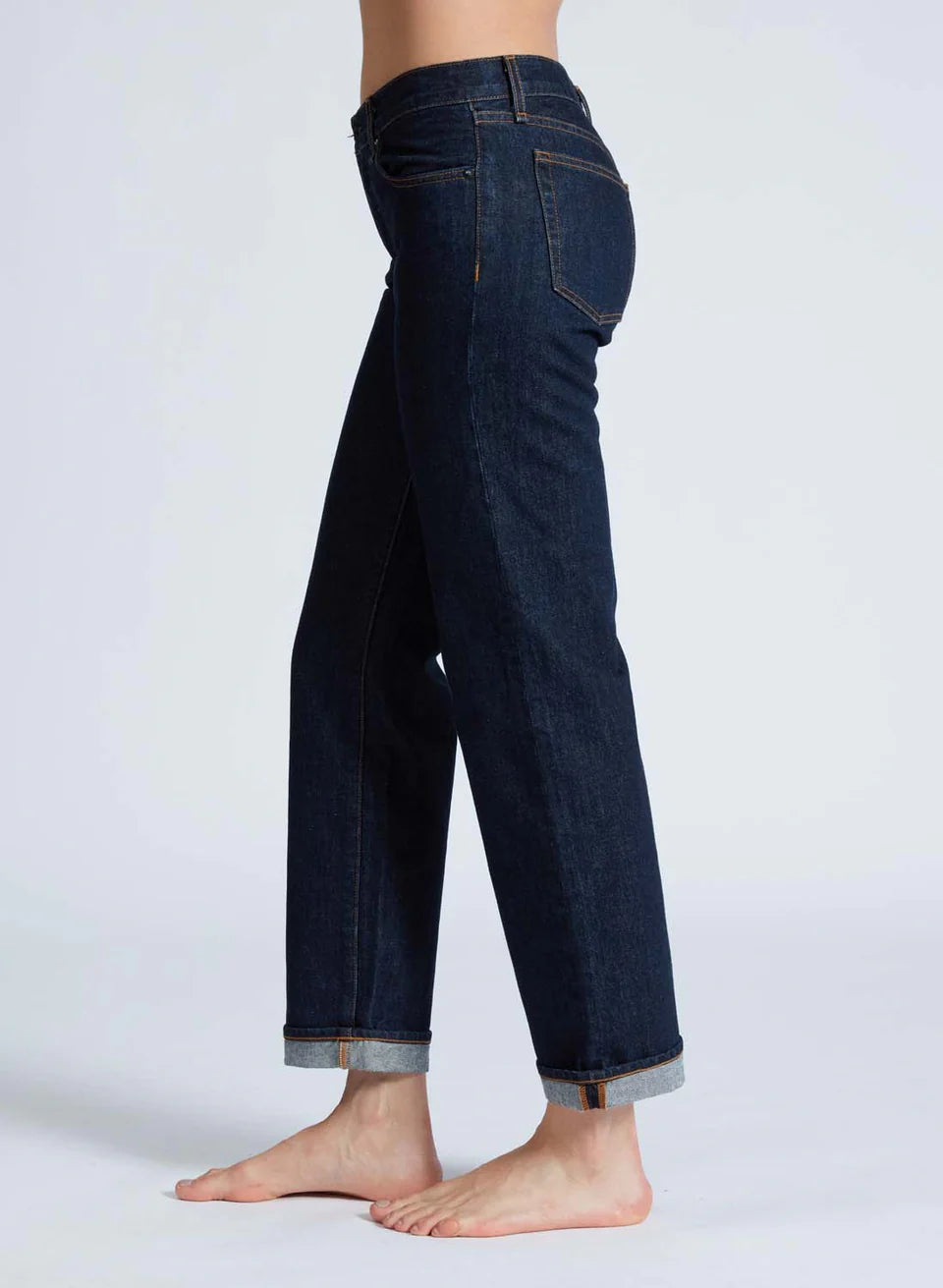 A side view of a person wearing ASKK's Low Rise Straight jeans in dark blue, crafted from Japanese denim with straight-leg cuffs. The barefoot individual stands against a light background, showcasing the 100% cotton jeans' classic five-pocket design and visible stitching for ultimate comfort.