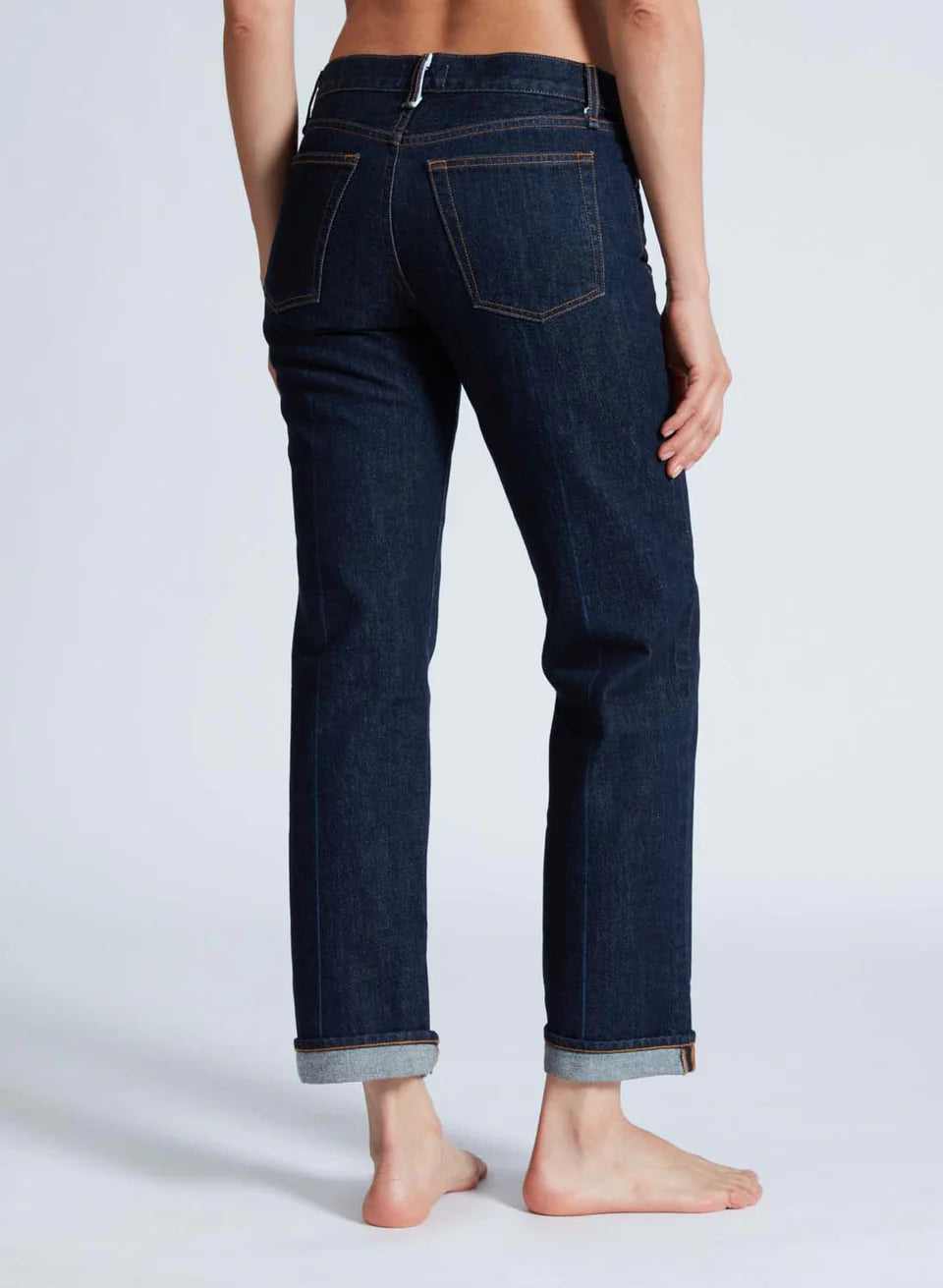 A person stands barefoot, wearing ASKK's Low Rise Straight jeans in dark blue. Crafted from 100% cotton Japanese denim with visible stitching and cuffed at the ankles, these jeans effortlessly complement the plain, light-colored background.