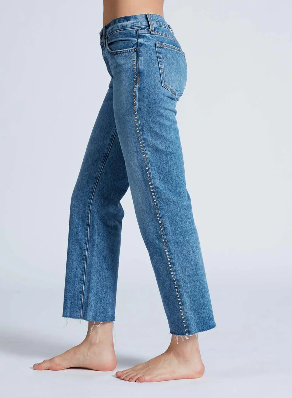 A person is barefoot on a light background, wearing the ASKK Low Rise Straight Studded jeans, crafted from raw hem Japanese denim in blue. The jeans have a straight cut with small decorative studs embellishing the side seams.