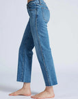 A person is barefoot on a light background, wearing the ASKK Low Rise Straight Studded jeans, crafted from raw hem Japanese denim in blue. The jeans have a straight cut with small decorative studs embellishing the side seams.