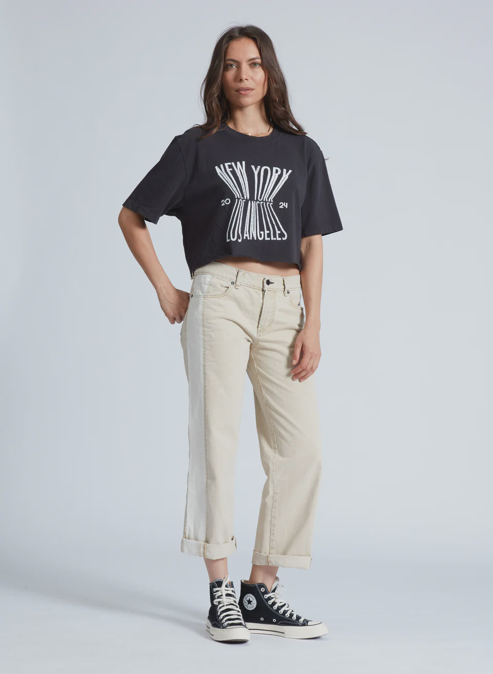 A person with long hair confidently wears the ASKK "Factory Girl" black T-shirt featuring "New York Los Angeles" alongside beige cuffed Japanese denim pants. Black high-top sneakers complete the look against a plain light background.