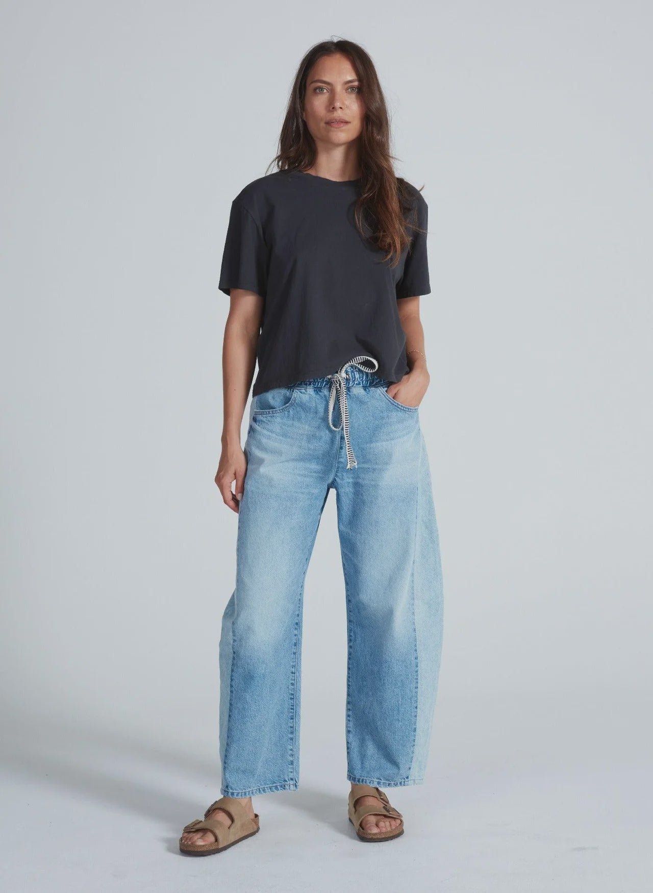 A woman with long brown hair and a relaxed expression stands against a plain background, wearing ASKK’s Cancun Pant—light blue Japanese denim jeans with wide legs and an elastic waistband—paired with a loose black t-shirt and tan sandals.