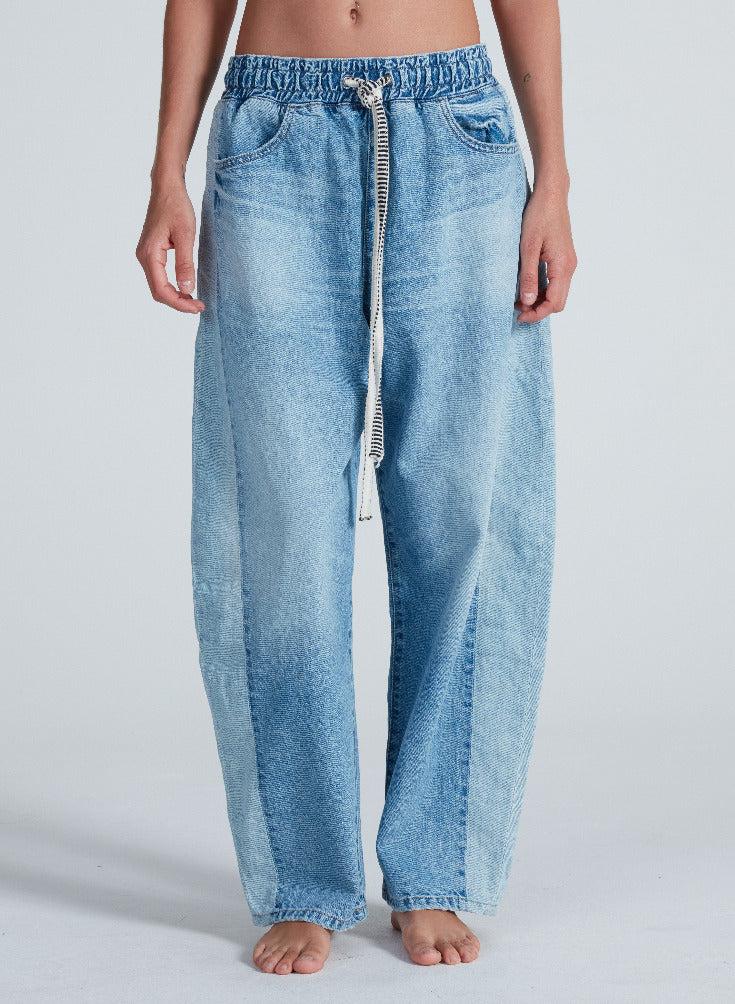 A person stands barefoot, hands by their sides, wearing the ASKK Cancun Pant—relaxed fit, light blue Japanese denim with an elastic waistband and drawstring—against a plain light background.
