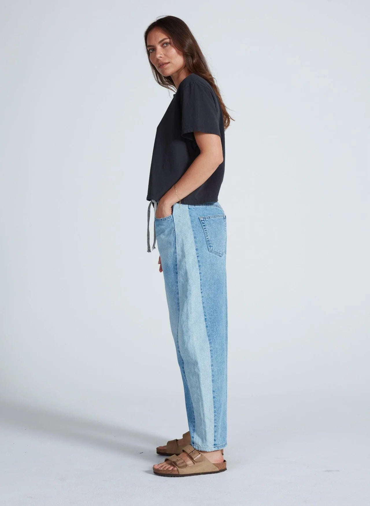 A woman stands sideways with her hands in her pockets, wearing ASKK’s Cancun Pant—relaxed fit two-tone blue jeans with an elastic waistband—a black t-shirt, and tan sandals. Her long hair is down against a light gray background.