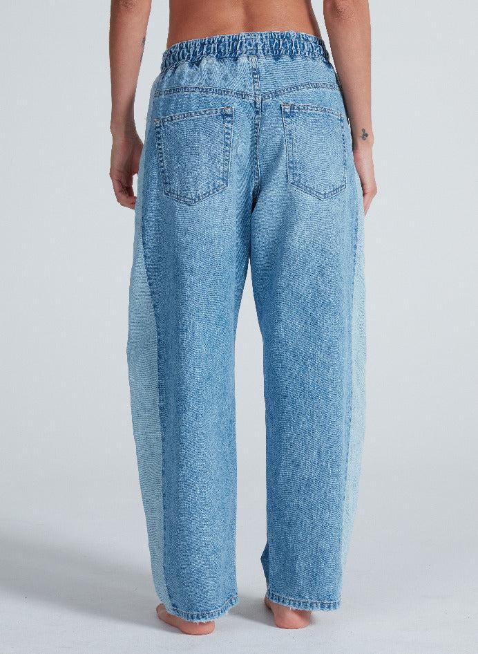 A person stands barefoot facing away, wearing light blue Cancun Pant jeans by ASKK with relaxed fit, back pockets, and an elastic waistband. The background is plain and light-colored.