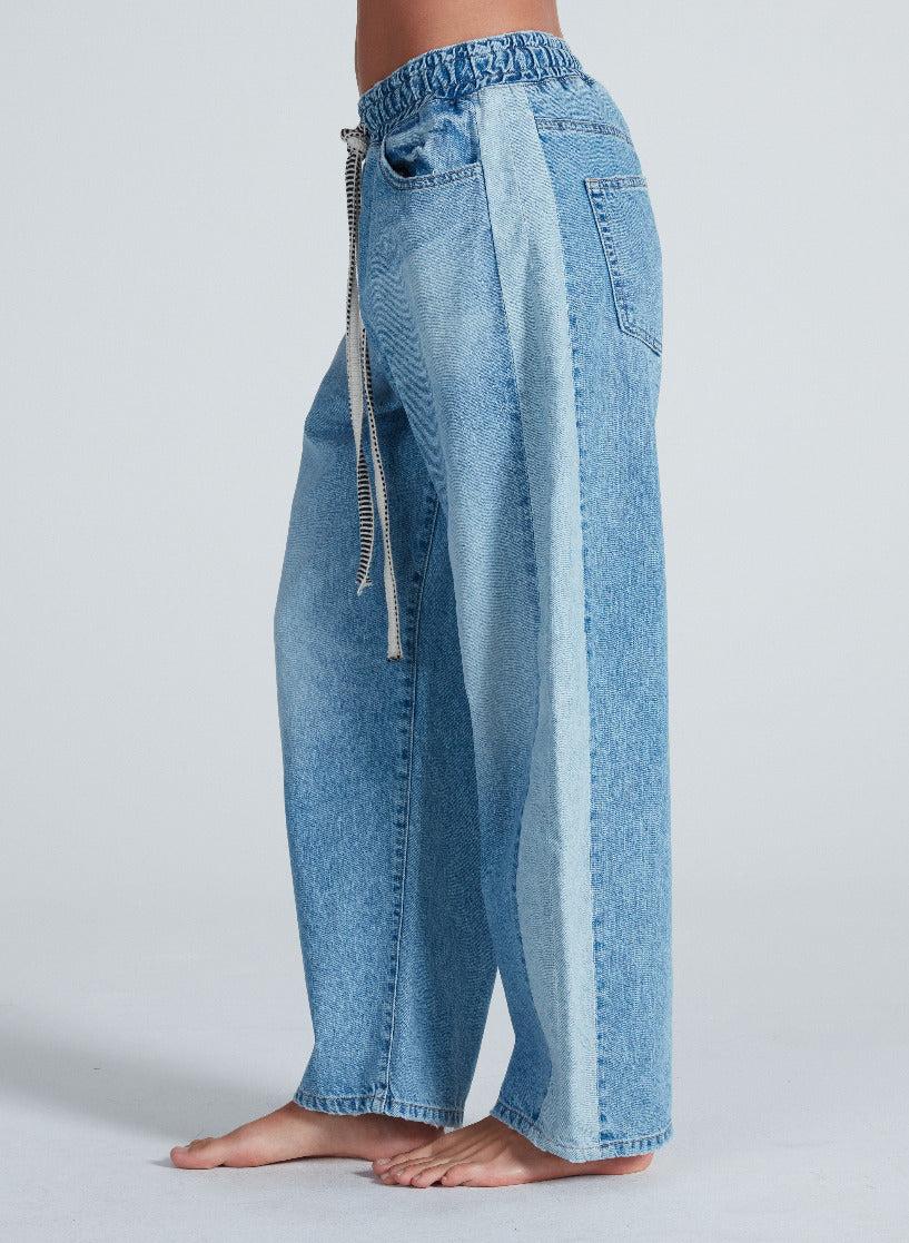 A person is wearing the ASKK Cancun Pant: light blue, wide-leg Japanese denim jeans with an elastic waistband, drawstring, and two-tone outer leg panels in a light Berkeley wash. The person is barefoot.
