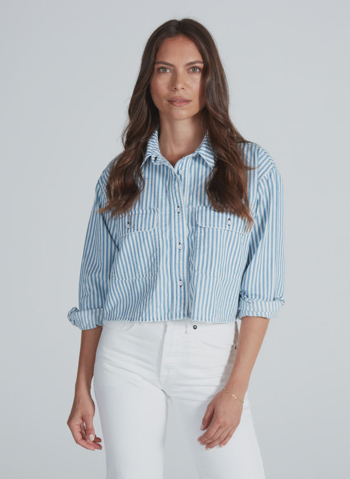 A woman with long brown hair wears the ASKK Cropped Nantucket Shirt—a blue and white vertically striped, lightweight cotton button-down—paired with high-waisted white pants against a plain light background.