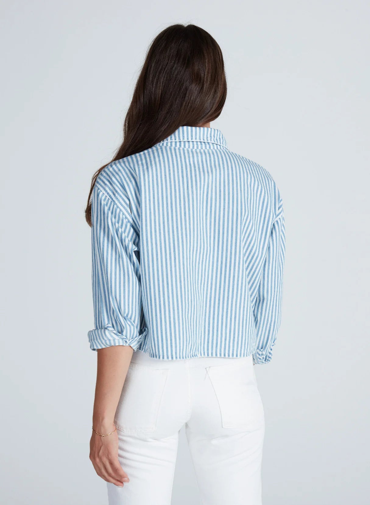 A woman with long brown hair, seen from behind, wears the ASKK Cropped Nantucket Shirt—blue and white striped Japanese shirting with double front pockets—paired with white pants, set against a light gray background.