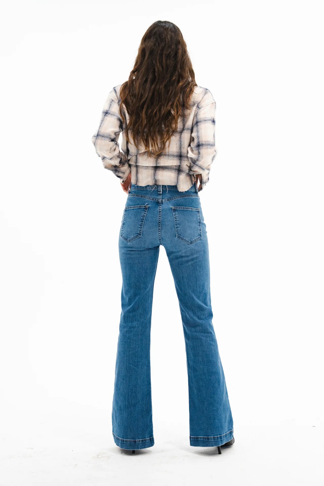 A woman with long wavy brown hair, wearing the ASKK Crosby Flannel in Japanese shirting fabric and high-waisted blue flared jeans, stands facing away against a plain white background, styled with black shoes.