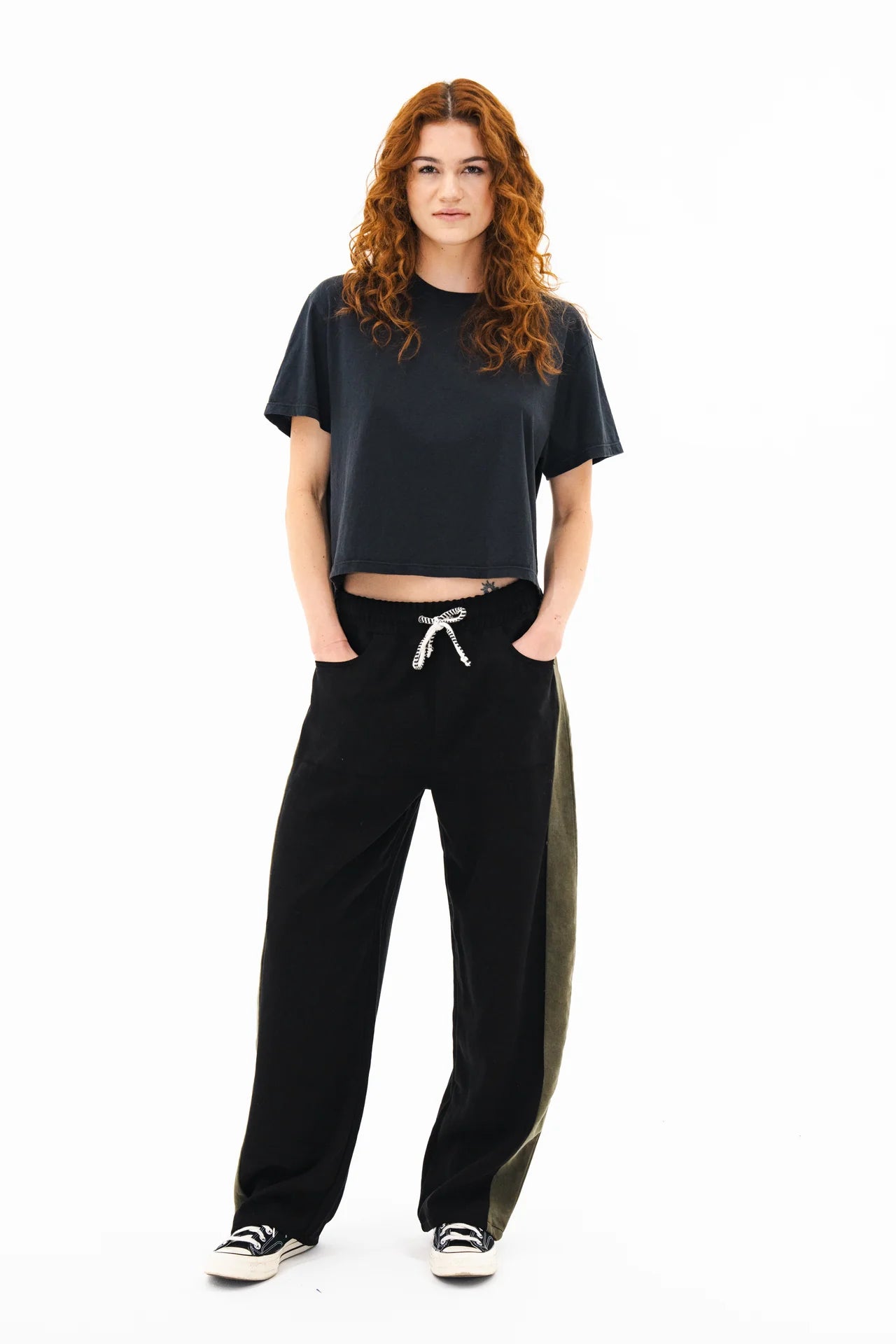 A woman with long curly red hair stands facing forward, wearing a loose dark t-shirt and ASKK’s Cancun Pant—wide leg, cropped drawstring black pants with an olive green side panel—plus black-and-white sneakers, hands in her pockets.
